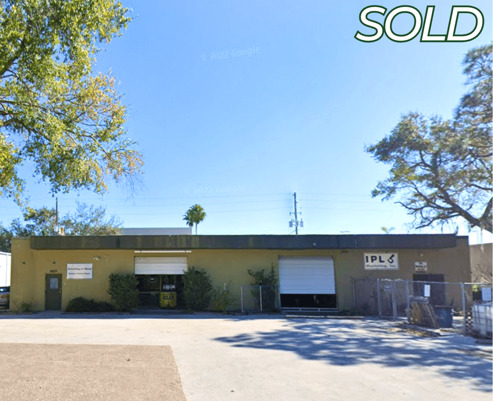 Single-story commercial property with two large garage doors, trees on both sides, and a "SOLD" label in the top right corner. Signs are visible on the front wall of this industrial commercial real estate.
