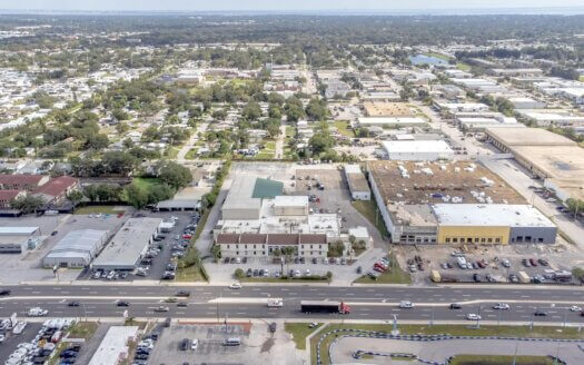 Aerial view of a suburban area featuring commercial real estate, parking lots, and a main road with several vehicles in motion. Residential houses and trees are visible in the background.