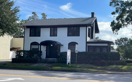A two-story white house with black trim and a dark roof, situated behind bushes on a sunny day with a clear sky. A street and crosswalk sign are visible in the foreground, suggesting potential as distinctive commercial property.