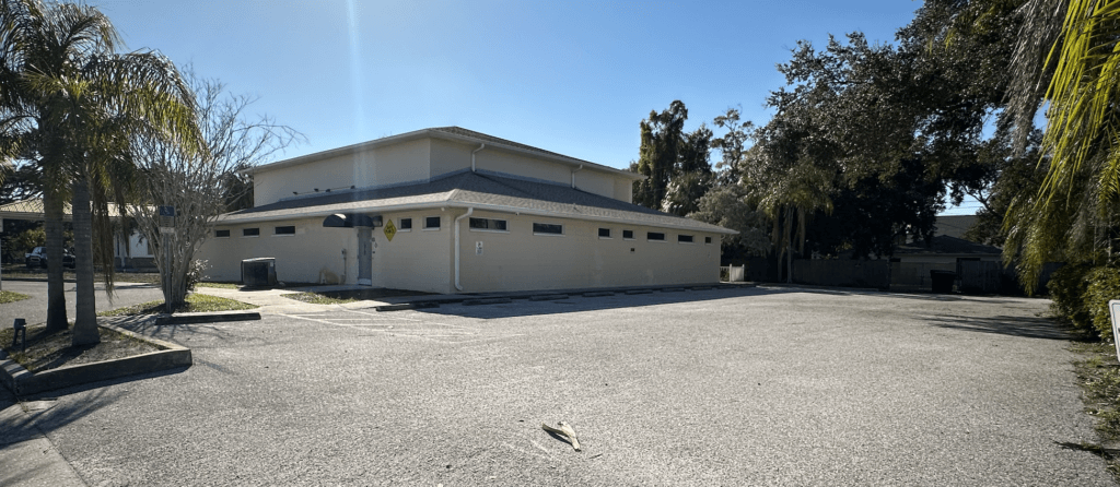A beige commercial property with a sloped roof sits adjacent to an empty parking lot under a clear blue sky, surrounded by palm and oak trees.