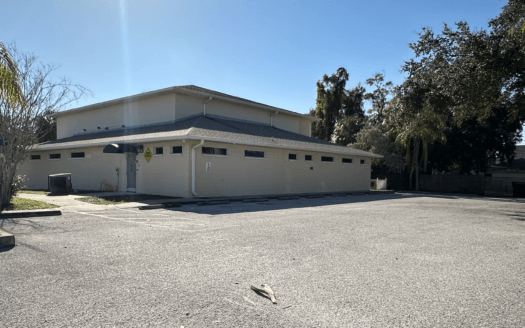A beige commercial property with a sloped roof sits adjacent to an empty parking lot under a clear blue sky, surrounded by palm and oak trees.
