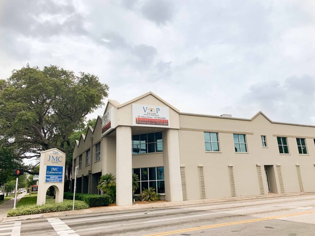 A two-story beige commercial property with a sign for JMC Contracting, located at a street corner with a large tree on the left and cloudy skies above.