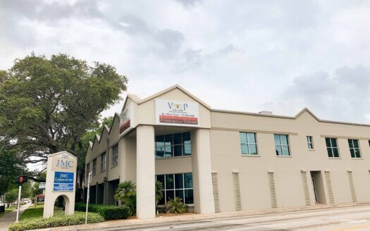A two-story beige commercial property with a sign for JMC Contracting, located at a street corner with a large tree on the left and cloudy skies above.