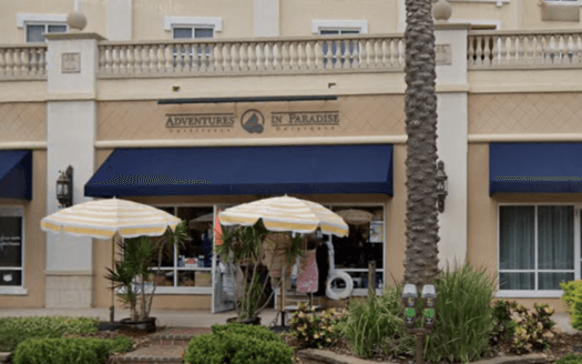 A storefront commercial property with blue awnings labeled "Adventures in Paradise" features two striped umbrellas, potted plants, and metered parking in front. The beige facade includes balconies above.
