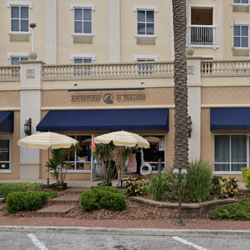A storefront commercial property with blue awnings labeled "Adventures in Paradise" features two striped umbrellas, potted plants, and metered parking in front. The beige facade includes balconies above.