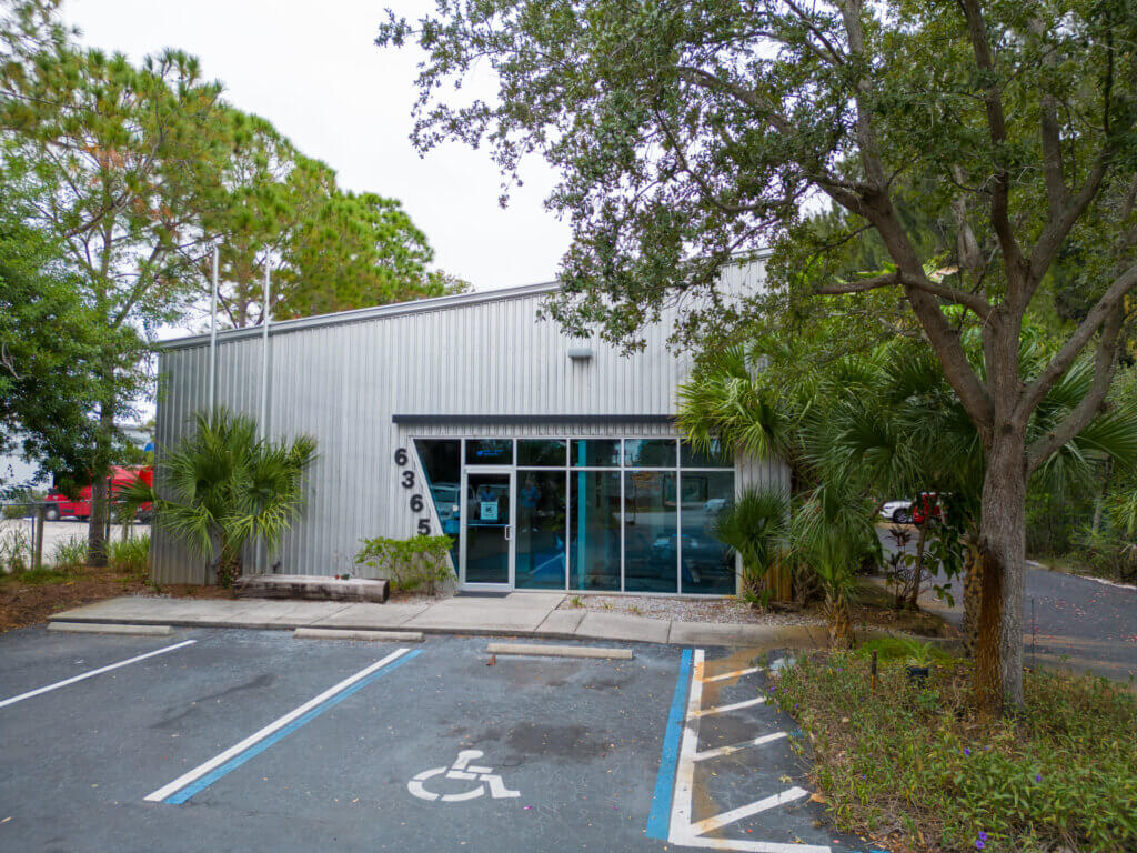 A small metal commercial property with a glass entrance, address number 636, and a handicap parking space in front, surrounded by trees and greenery.
