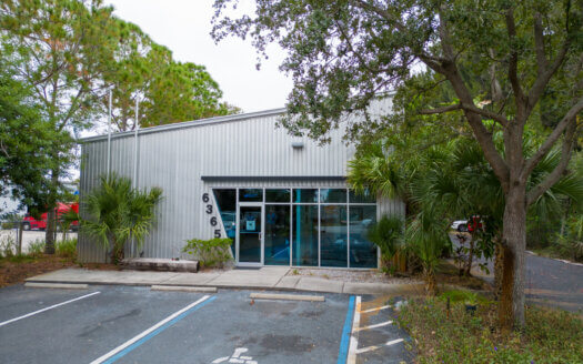 A small metal commercial property with a glass entrance, address number 636, and a handicap parking space in front, surrounded by trees and greenery.