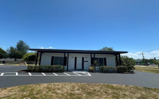 A small, single-story white commercial property with dark trim, surrounded by shrubs and parking spaces, under a clear blue sky.