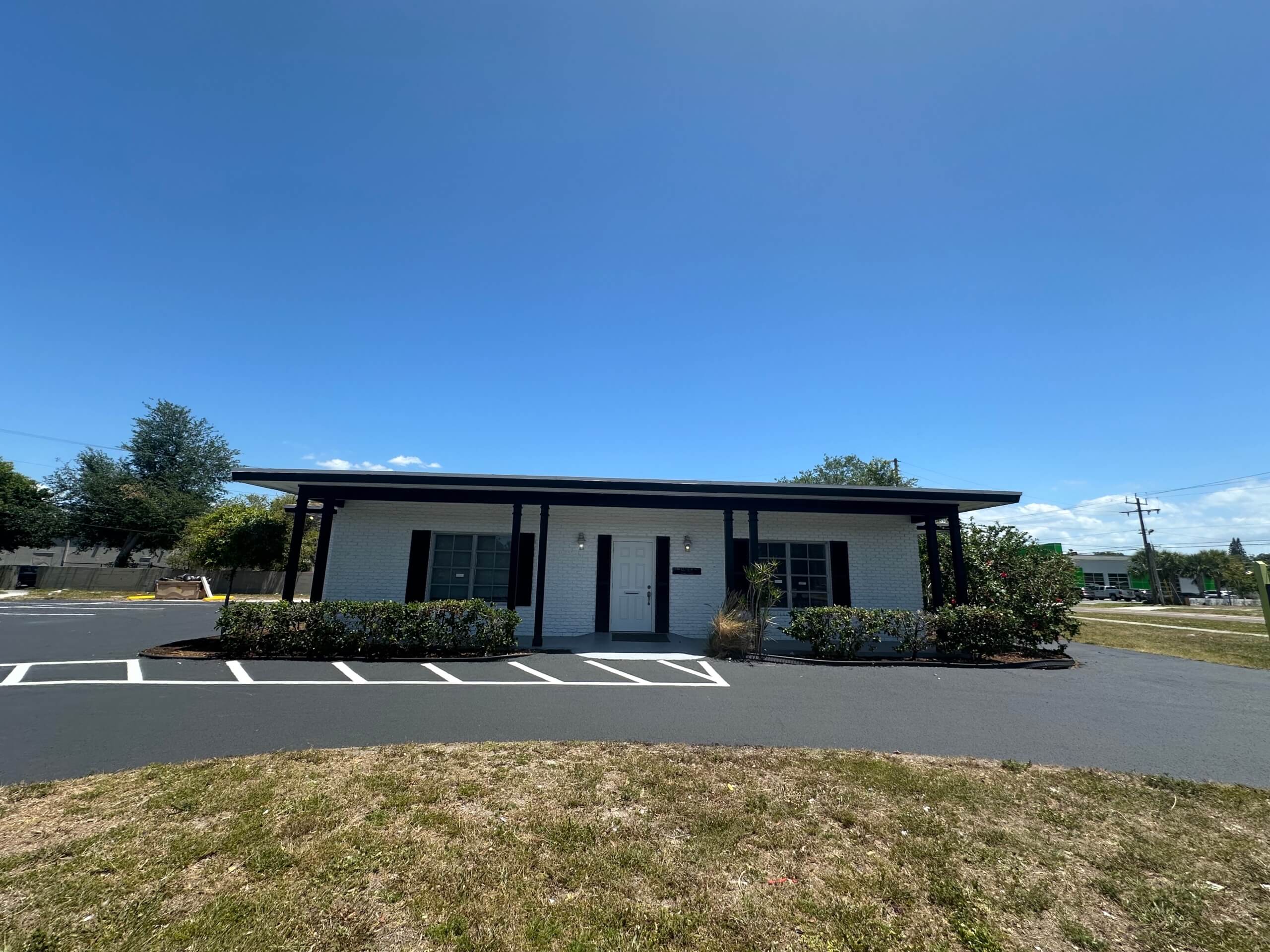 A small, single-story white commercial property with dark trim, surrounded by shrubs and parking spaces, under a clear blue sky.