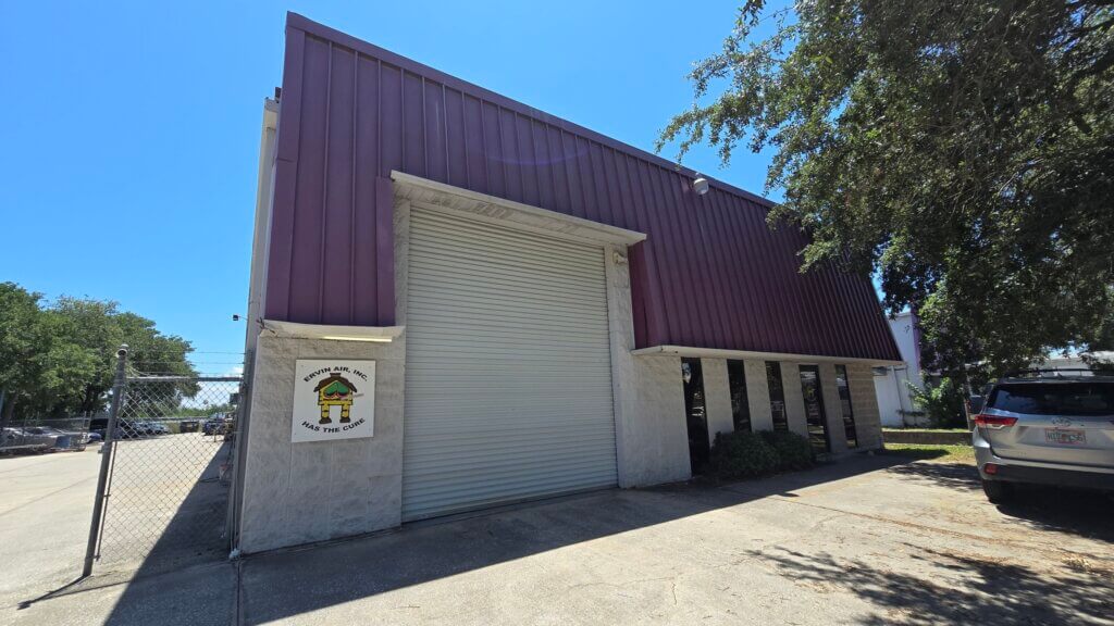 A warehouse-style commercial property with a purple roof, white exterior walls, a large closed roll-up door, and a sign next to the entrance. A parked car and tree are visible on the right.