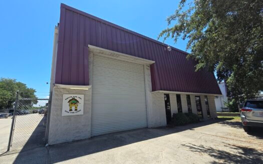 A warehouse-style commercial property with a purple roof, white exterior walls, a large closed roll-up door, and a sign next to the entrance. A parked car and tree are visible on the right.