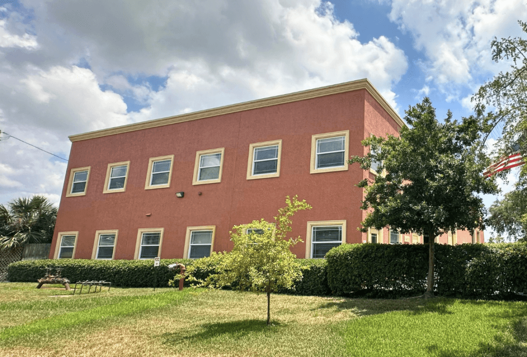A two-story red commercial property with multiple windows, surrounded by green grass, hedges, a small tree, and an American flag on the right side under a partly cloudy sky.