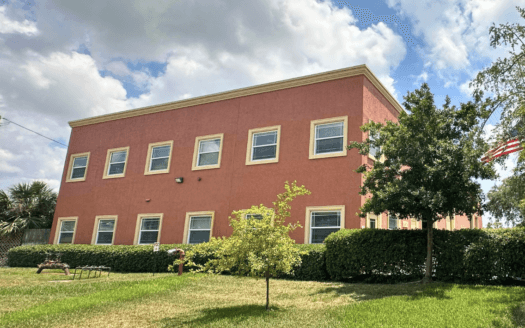 A two-story red commercial property with multiple windows, surrounded by green grass, hedges, a small tree, and an American flag on the right side under a partly cloudy sky.