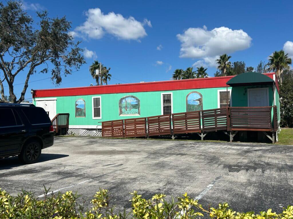 A green and red single-story commercial property with arched windows and a wooden accessibility ramp, set in a sunny parking lot with palm trees in the background.