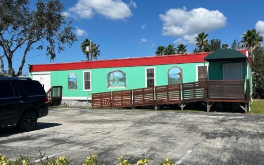 A green and red single-story commercial property with arched windows and a wooden accessibility ramp, set in a sunny parking lot with palm trees in the background.