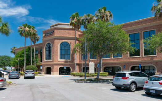 A modern brick commercial property with large arched windows, surrounded by palm trees and parked cars under a clear blue sky.