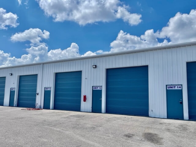 Row of white self-storage units with blue roll-up doors and unit numbers 12, 13, and 14 visible under a partly cloudy sky—ideal commercial property for your storage or commercial real estate needs.