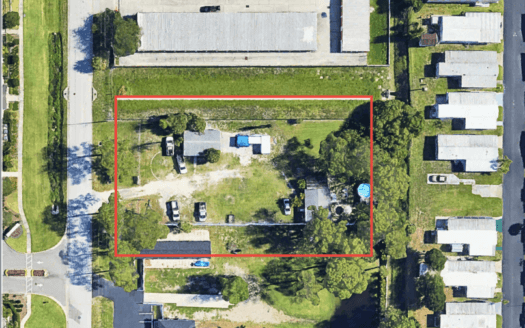 Aerial view of a rectangular commercial property outlined in red with scattered vehicles and trees, surrounded by buildings and houses; "SOLD" is labeled in the top right corner.