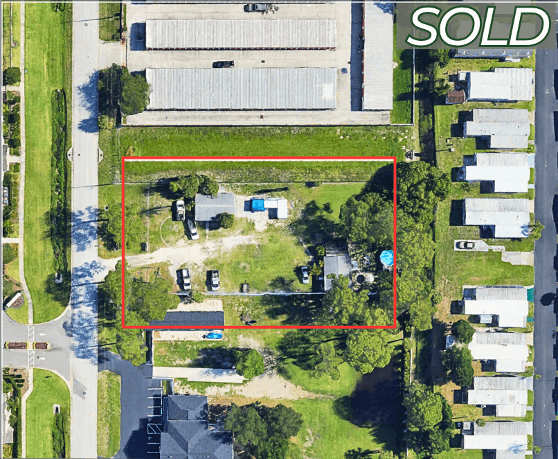 Aerial view of a rectangular commercial property outlined in red with scattered vehicles and trees, surrounded by buildings and houses; "SOLD" is labeled in the top right corner.