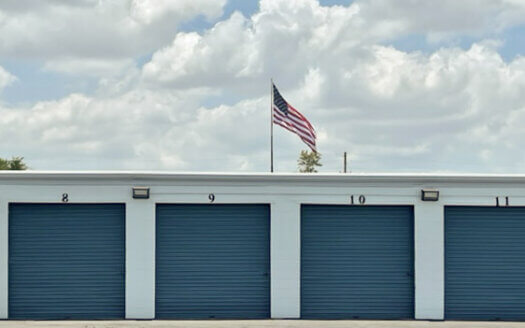 Four blue storage unit doors numbered 8 to 11 on a white commercial property, with an American flag on a pole above the roof against a cloudy sky.