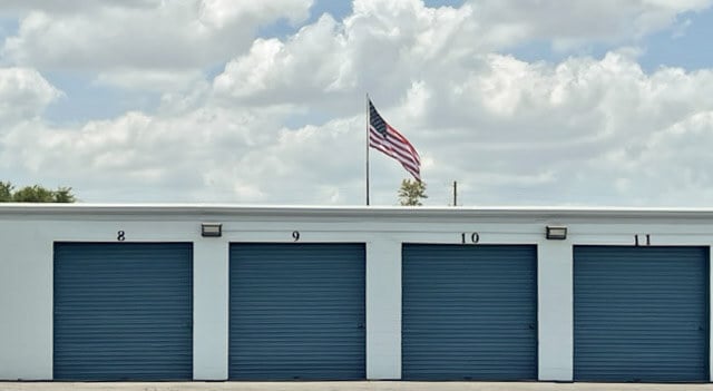 Four blue storage unit doors numbered 8 to 11 on a white commercial property, with an American flag on a pole above the roof against a cloudy sky.