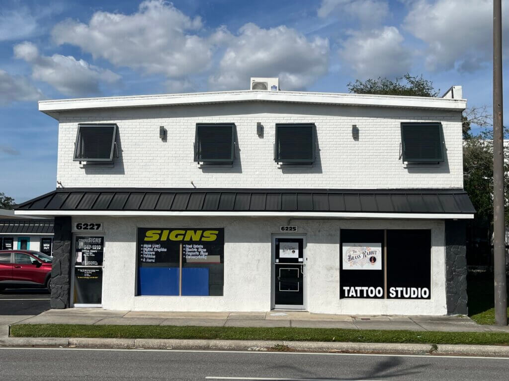 A white two-story commercial property with black trim houses a signs shop and a tattoo studio. Signs and business names are displayed on the windows and doors.