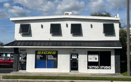 A white two-story commercial property with black trim houses a signs shop and a tattoo studio. Signs and business names are displayed on the windows and doors.