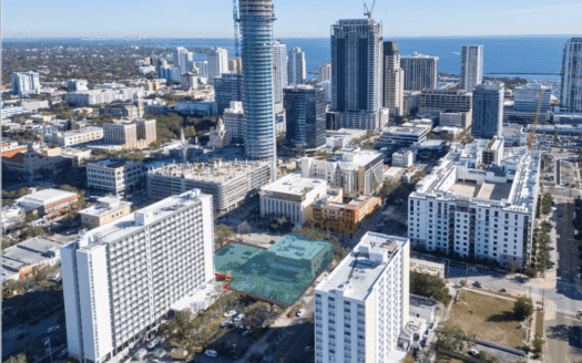 Aerial view of a cityscape with high-rise buildings and commercial property, including a tall tower under construction with cranes, near the coastline on a clear day.