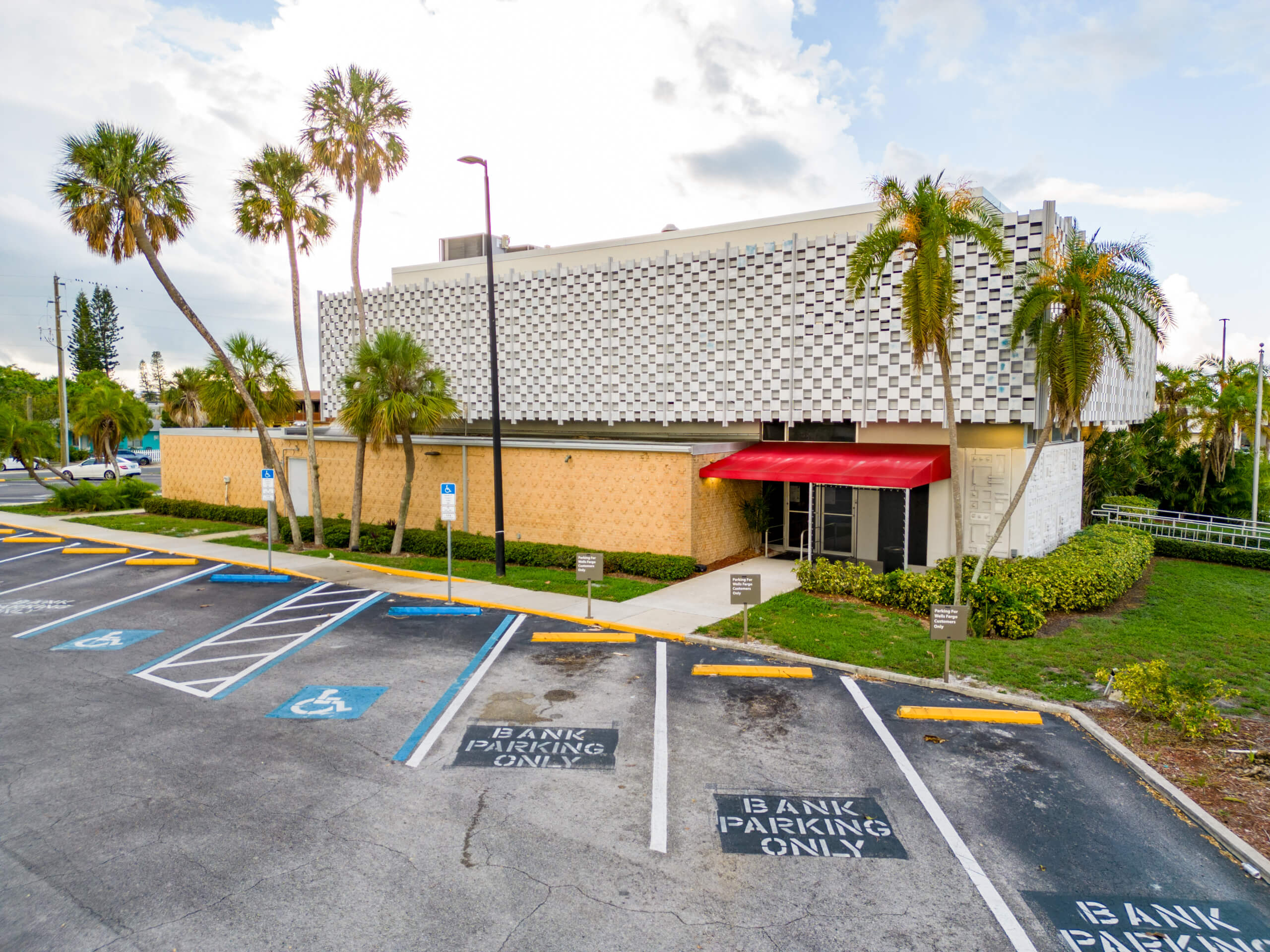 A commercial property with a patterned exterior, red awning, and palm trees in front, featuring parking spaces labeled for bank use and accessible parking.