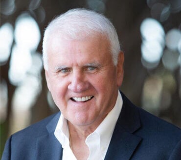 Kenneth Heretick, an older man with short white hair, wearing a dark blazer and white shirt, smiles at the camera in an outdoor setting—a fitting portrait for a professional in vector commercial real estate.