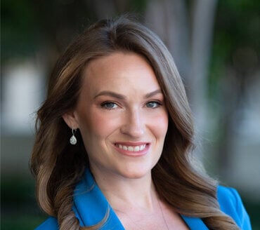 Ashley Willis, a woman with long light brown hair, wears a blue blazer and earrings, smiling at the camera against a blurred outdoor background.