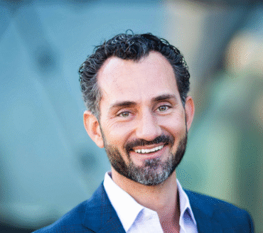 Kenneth Heretick, a man with dark curly hair and a beard, wears a blue blazer and white shirt, smiling at the camera with a blurred background.