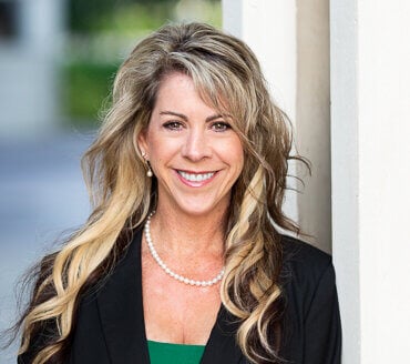Erica Delgado, a woman with long blonde hair, stands smiling in front of a light-colored wall, dressed in a black blazer, green top, and pearl necklace.
