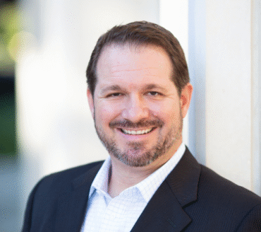 Michael Ludwig, a man with short brown hair and beard, is smiling while wearing a dark suit jacket and white checkered shirt, standing by a light-colored wall.