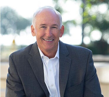 A middle-aged man with short gray hair in a dark suit jacket and white shirt smiles while seated outdoors.