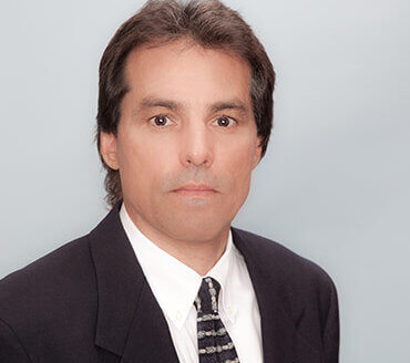 A man with straight brown hair, wearing a dark suit jacket, white dress shirt, and patterned tie, poses against a plain light background—epitomizing the classic style of montana massa.