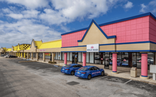 A strip mall with colorful storefronts, mostly pink and yellow, sits along US HWY 19 in Clearwater, featuring leased units like Molly Maid and two branded blue cars parked out front under a partly cloudy sky.