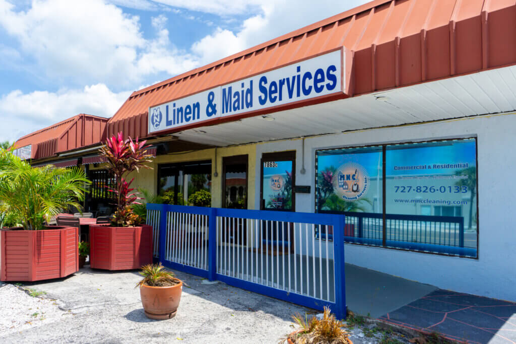 A storefront commercial property with a sign reading "Linen & Maid Services" and large windows displaying business information, located under a brown awning with potted plants outside.