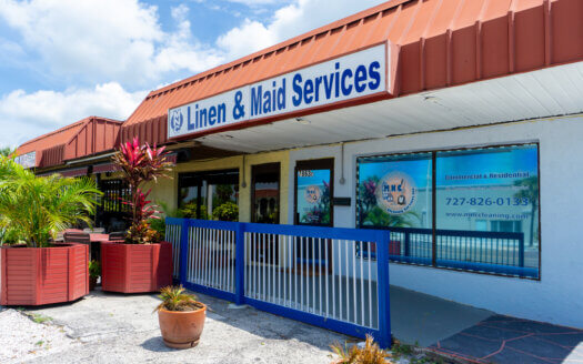 A storefront commercial property with a sign reading "Linen & Maid Services" and large windows displaying business information, located under a brown awning with potted plants outside.