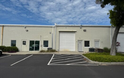 Single-story commercial property featuring a closed white roll-up garage door, two regular doors, and empty parking spaces out front beneath a partly cloudy sky.