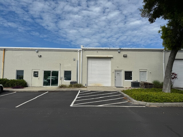 Single-story commercial property featuring a closed white roll-up garage door, two regular doors, and empty parking spaces out front beneath a partly cloudy sky.