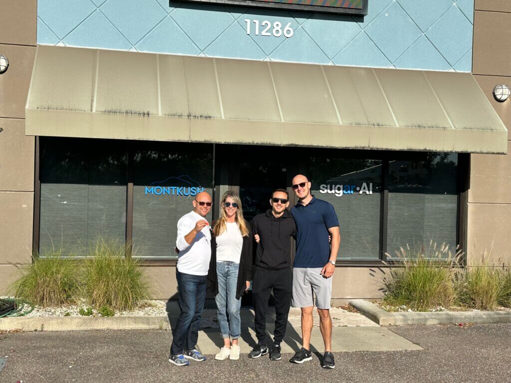 Four people pose under a tan awning in front of a building with address 11286 on US HWY 19 Clearwater; two glass doors read "MONTKUSH" and "sugar AI," highlighting these leased property Clearwater commercial units.