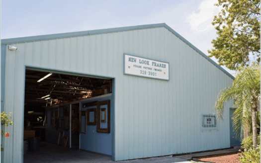 A light blue industrial building, ideal as commercial property, features an open garage door and a “New Look Frames” sign. Framed items are visible inside, with trees standing to the right of the building.