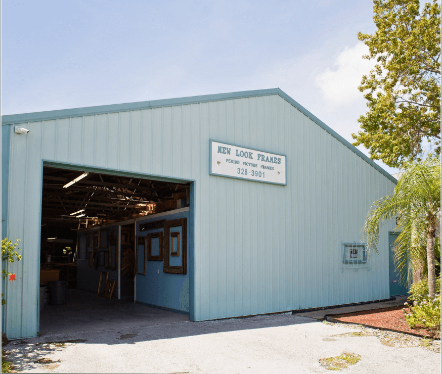 A light blue industrial building, ideal as commercial property, features an open garage door and a “New Look Frames” sign. Framed items are visible inside, with trees standing to the right of the building.