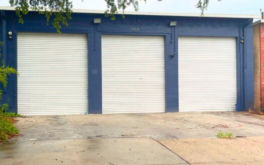 Three closed white garage doors are set in a blue commercial property with the number 2464 above the middle door. A tree branch partially covers the top of the image.