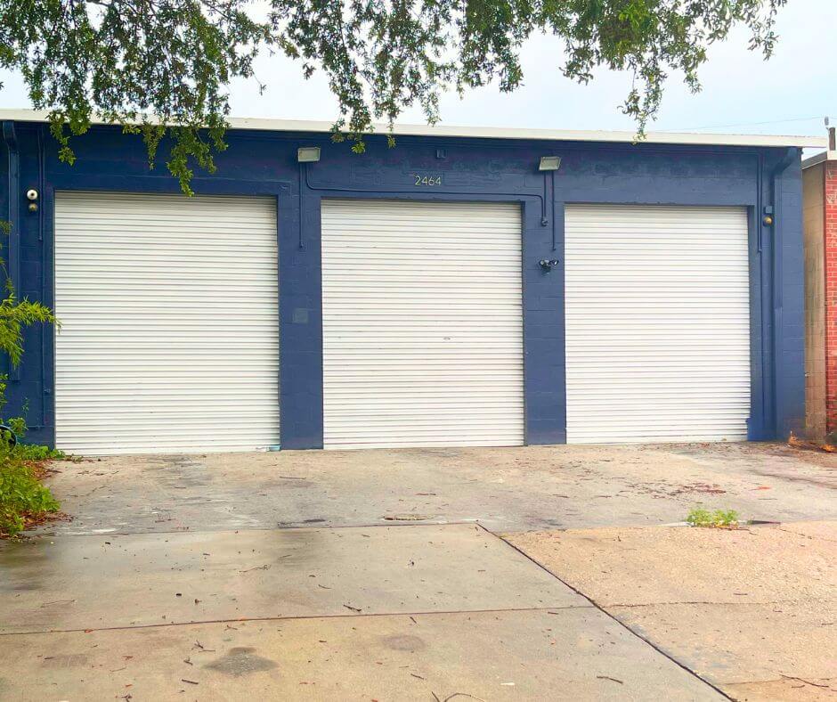 Three closed white garage doors are set in a blue commercial property with the number 2464 above the middle door. A tree branch partially covers the top of the image.