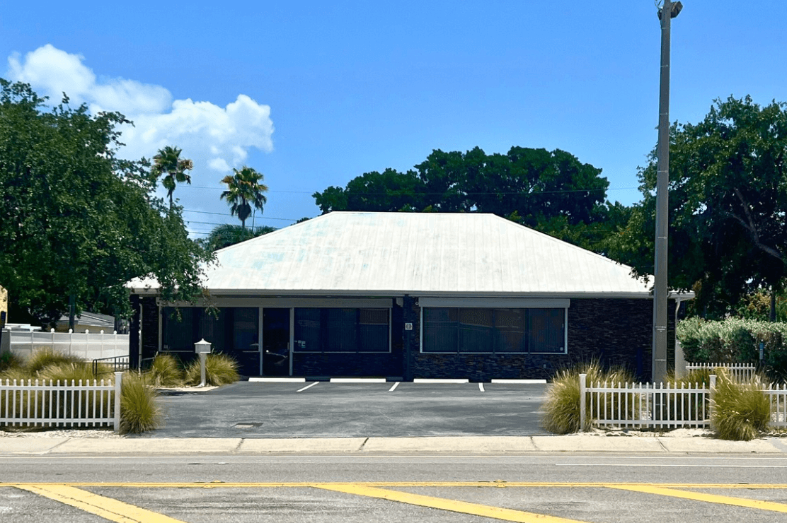 A small, one-story commercial property with a white roof, large front windows, and a picket fence sits in front of a few parking spaces on a sunny day.