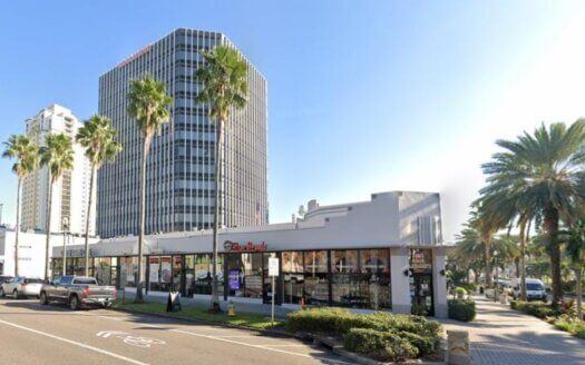 A single-story commercial property with large windows sits in front of a tall office tower and palm trees on a sunny city street. Cars are parked along the curb.