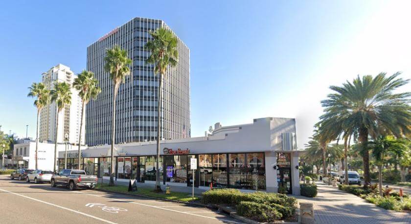 A single-story commercial property with large windows sits in front of a tall office tower and palm trees on a sunny city street. Cars are parked along the curb.