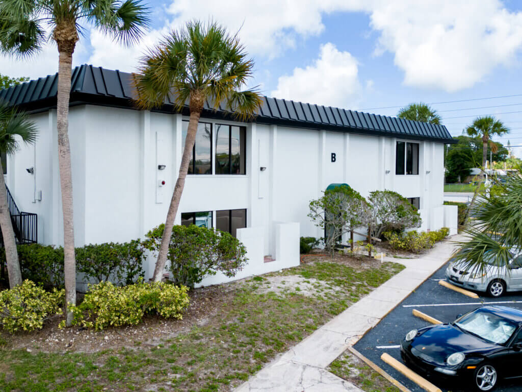 Two-story white commercial property with black roof and windows, surrounded by palm trees and shrubs, with parked cars in a partially visible parking lot.
